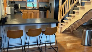 A modern kitchen with wooden cabinets, a stainless steel refrigerator, a bar area with stools, and a wooden staircase.