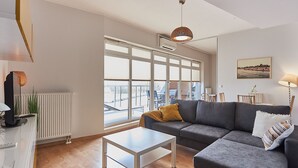 Una sala de estar moderna con un sofá gris, una mesa de centro blanca y ventanas amplias que ofrecen vistas de la ciudad.