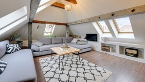 Uma sala de estar estilo loft com um sofá cinzento, uma mesa de centro de madeira e um tapete estampado. Há claraboias, uma televisão e uma estante com diversos objetos.