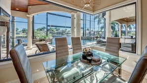 Uma ampla área de refeições com mesa e cadeiras de vidro, janelas grandes com vista para o exterior e um lustre moderno.