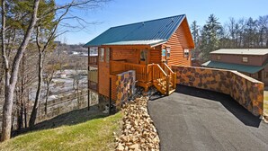 A wooden cabin with a stone retaining wall, a metal roof, and a view of a parking area and trees. - Mountain and City light views! Close to Strip, Private Indoor Pool + Free Tix (Pigeon Forge)