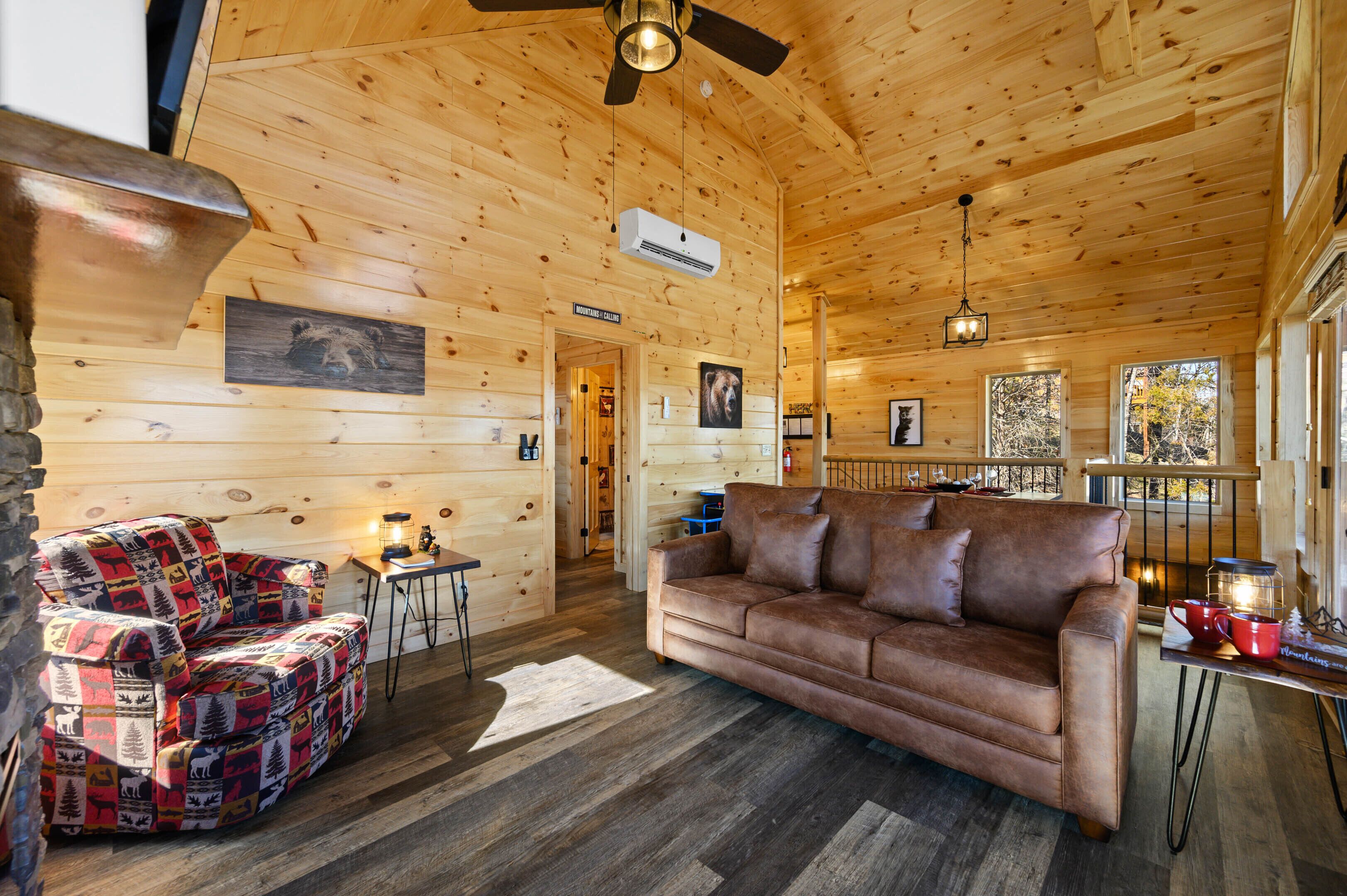 A cozy cabin interior with a wooden ceiling, a brown leather sofa, a colorful quilted chair, and a stone wall.
