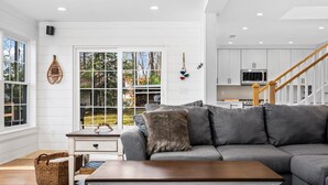 Ein modernes Wohnzimmer mit einem grauen Ecksofa, einem Holztisch und Blick ins Freie durch große Fenster.