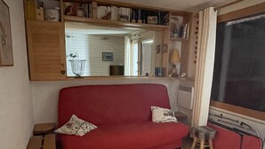 Un sofà vermell amb dos coixins, un tamboret petit, un armari de fusta i un mirall a la paret.