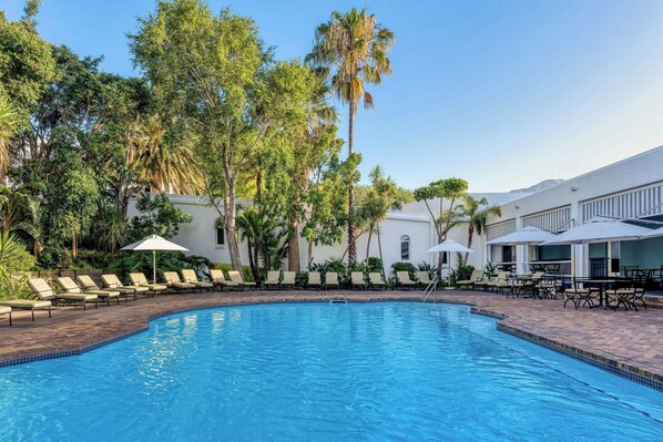 Una piscina con sillones de descanso y sombrillas, rodeada de palmeras y un edificio con terraza.