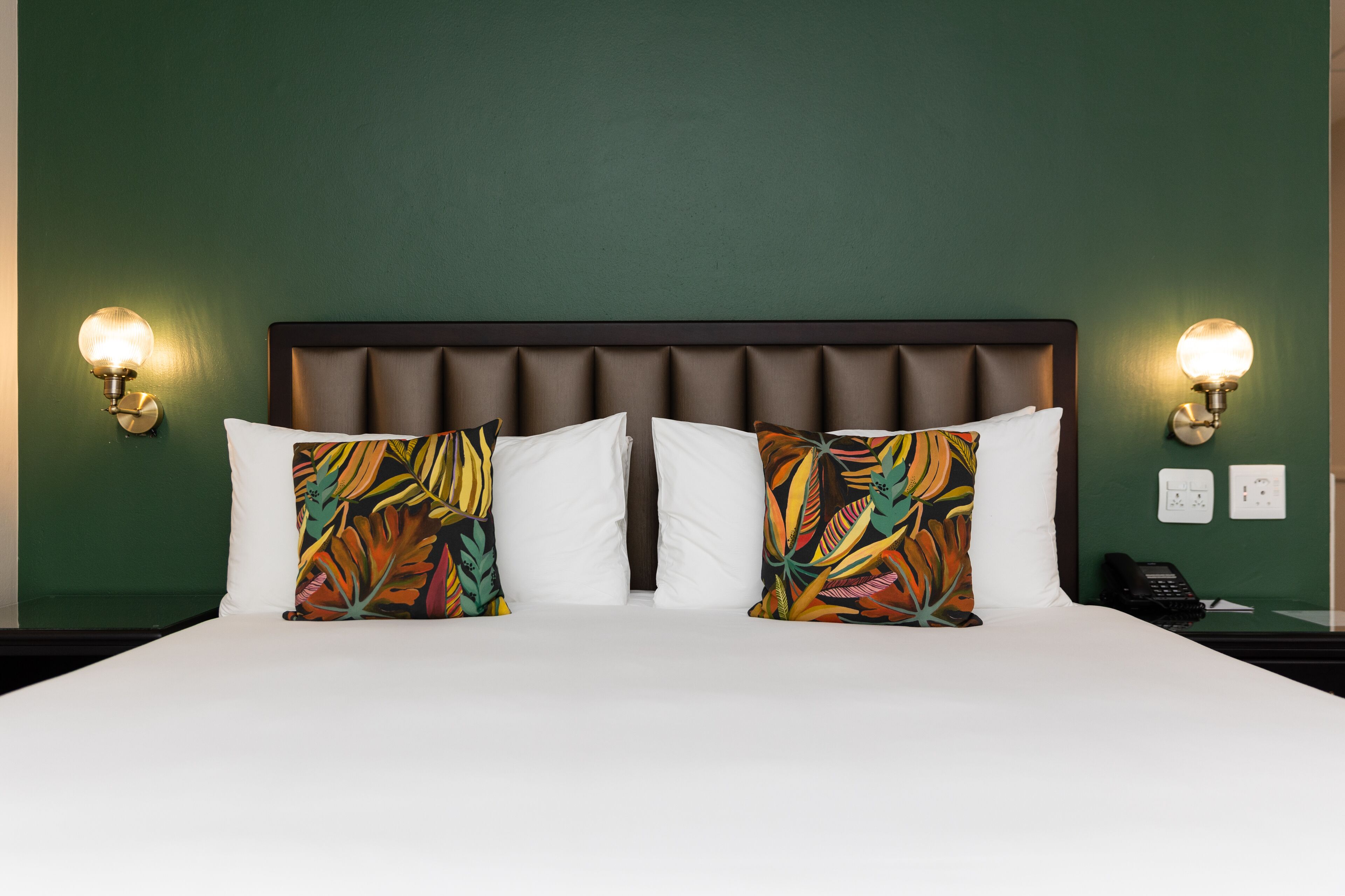 Habitación de hotel con una cama grande, dos almohadones decorativos, un cabecero y luces fijadas a la pared.