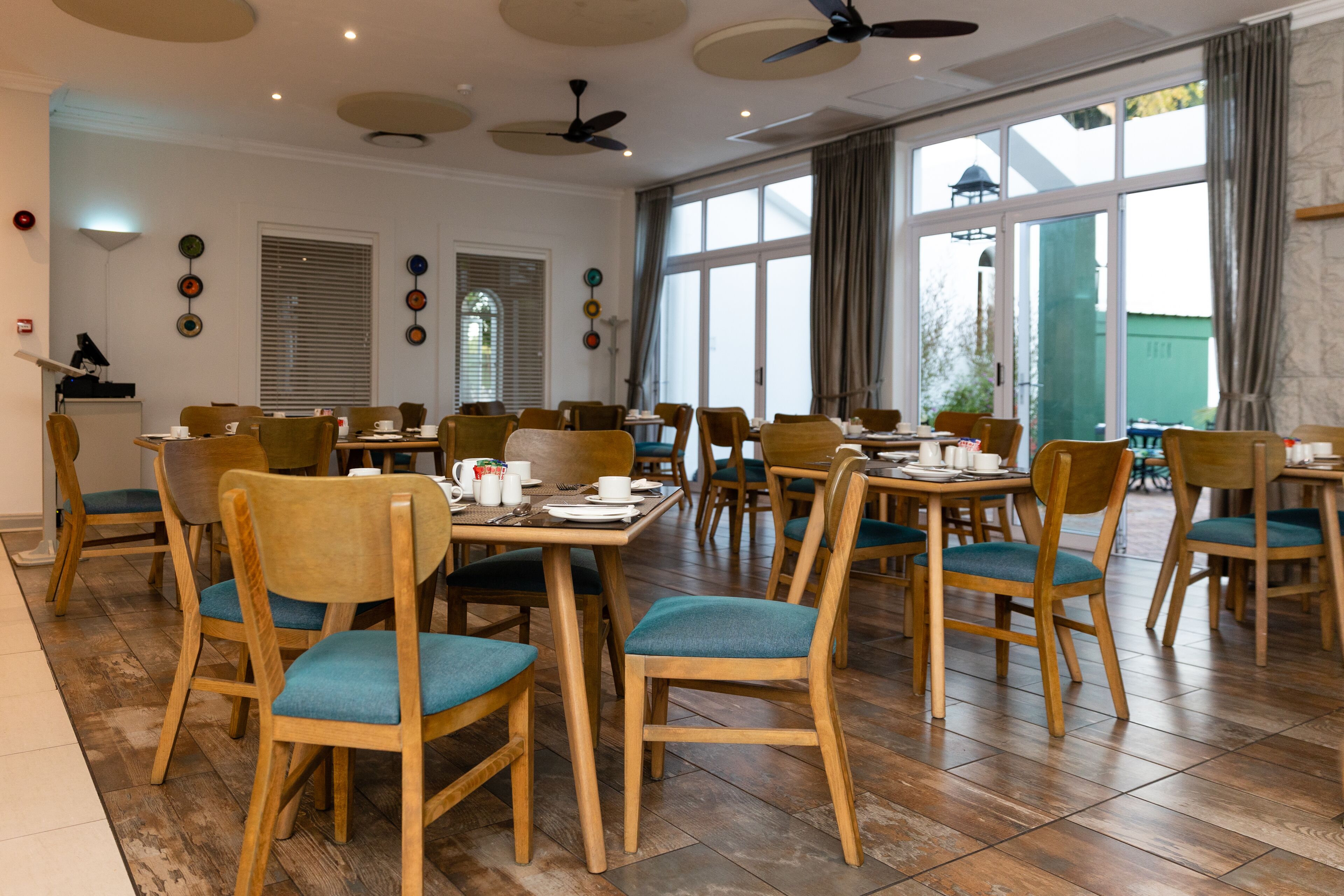 Un vaste espace dédié à la salle à manger, avec des chaises et des tables en bois, de grandes fenêtres dotées de rideaux et des ventilateurs de plafond.