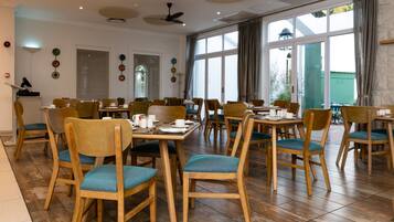 A spacious dining area with wooden chairs and tables, large windows with curtains, and ceiling fans.