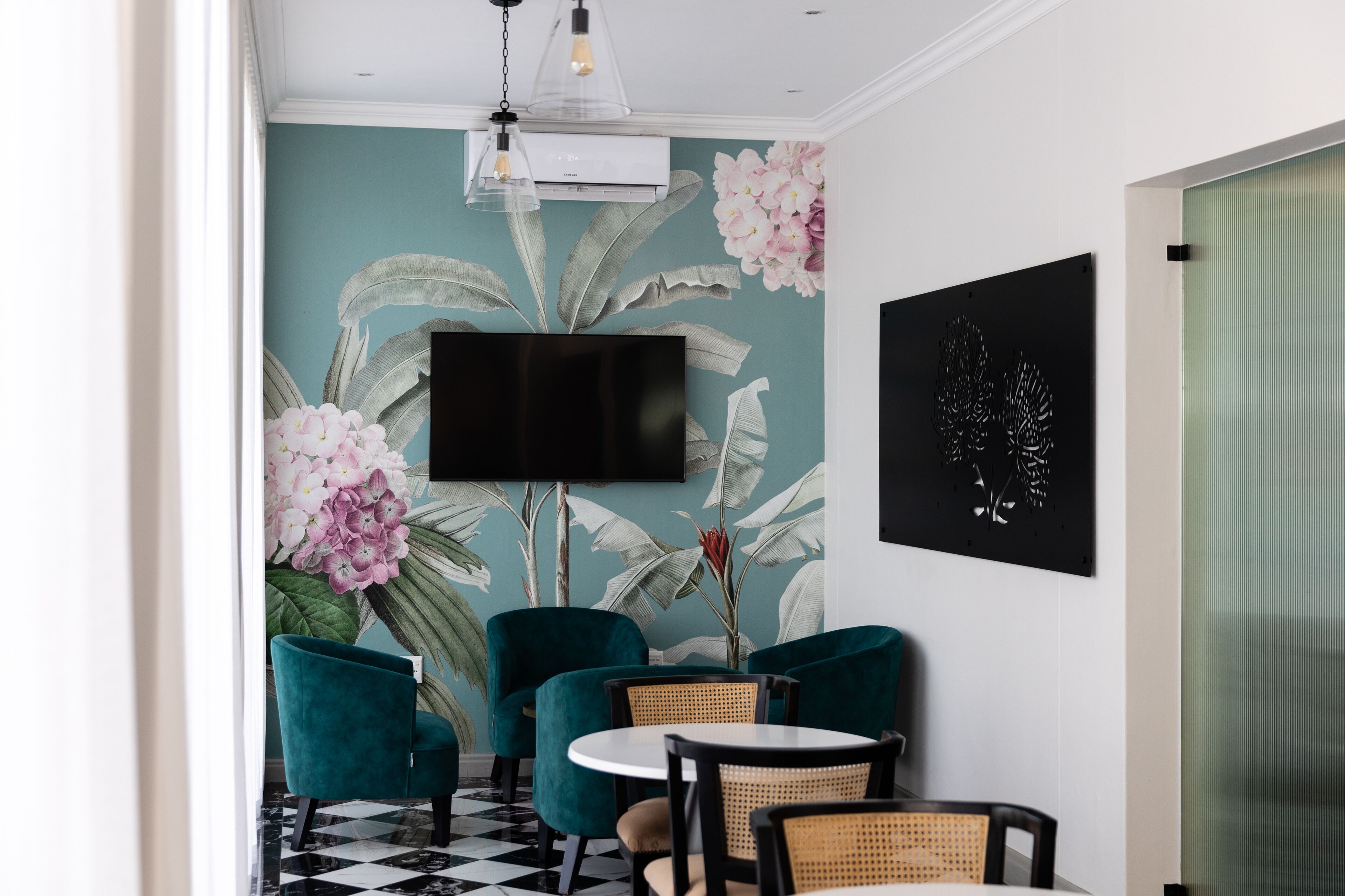 A room with teal walls, a large floral mural, teal chairs, a round table, a flat-screen TV, and a modern light fixture.
