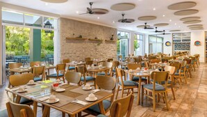 Un comedor amplio con varias mesas y sillas, ventiladores de techo y ventanas grandes que ofrecen vistas al exterior.