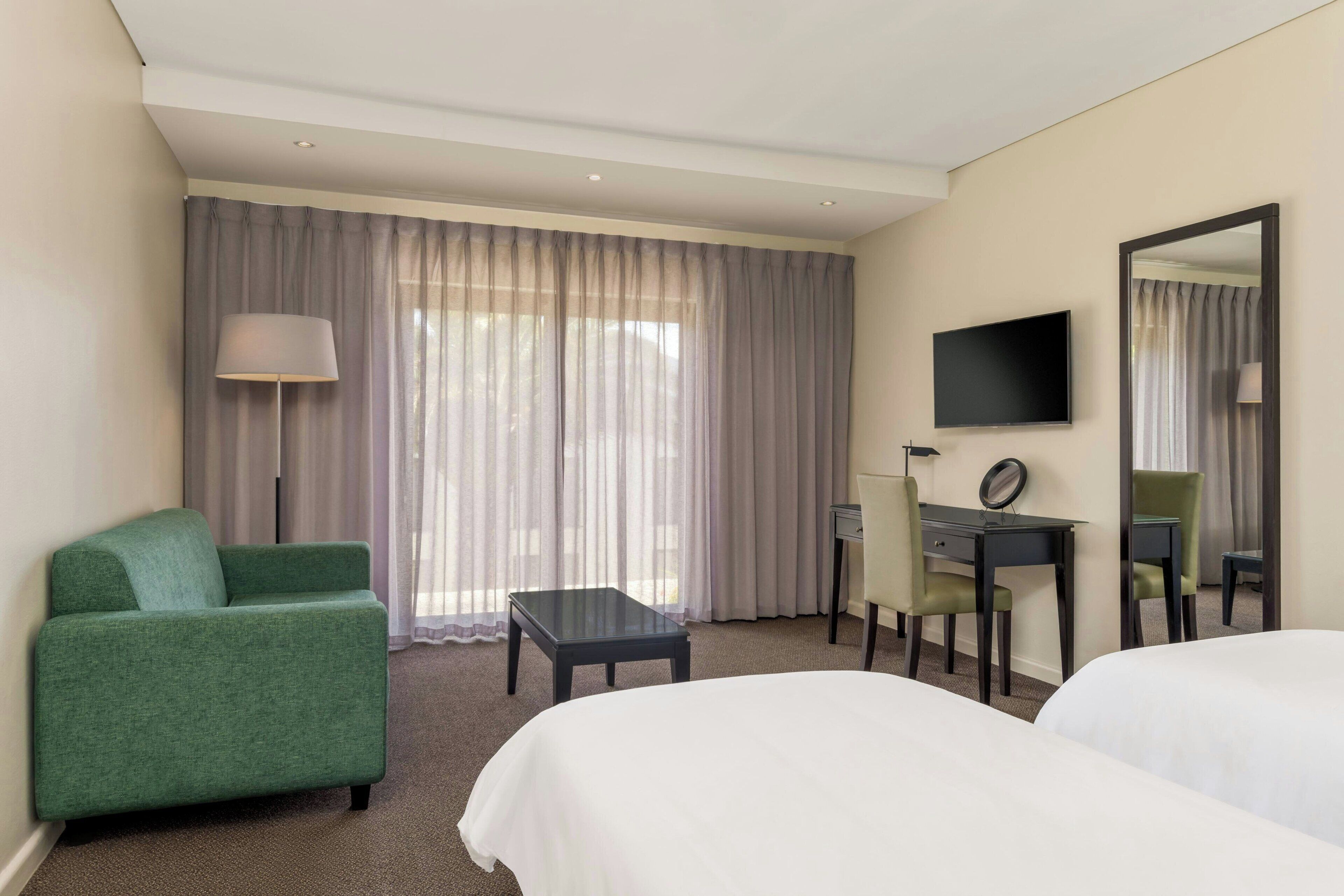 A hotel room with two beds, a desk with a chair, a TV, a lamp, and a large window with curtains.