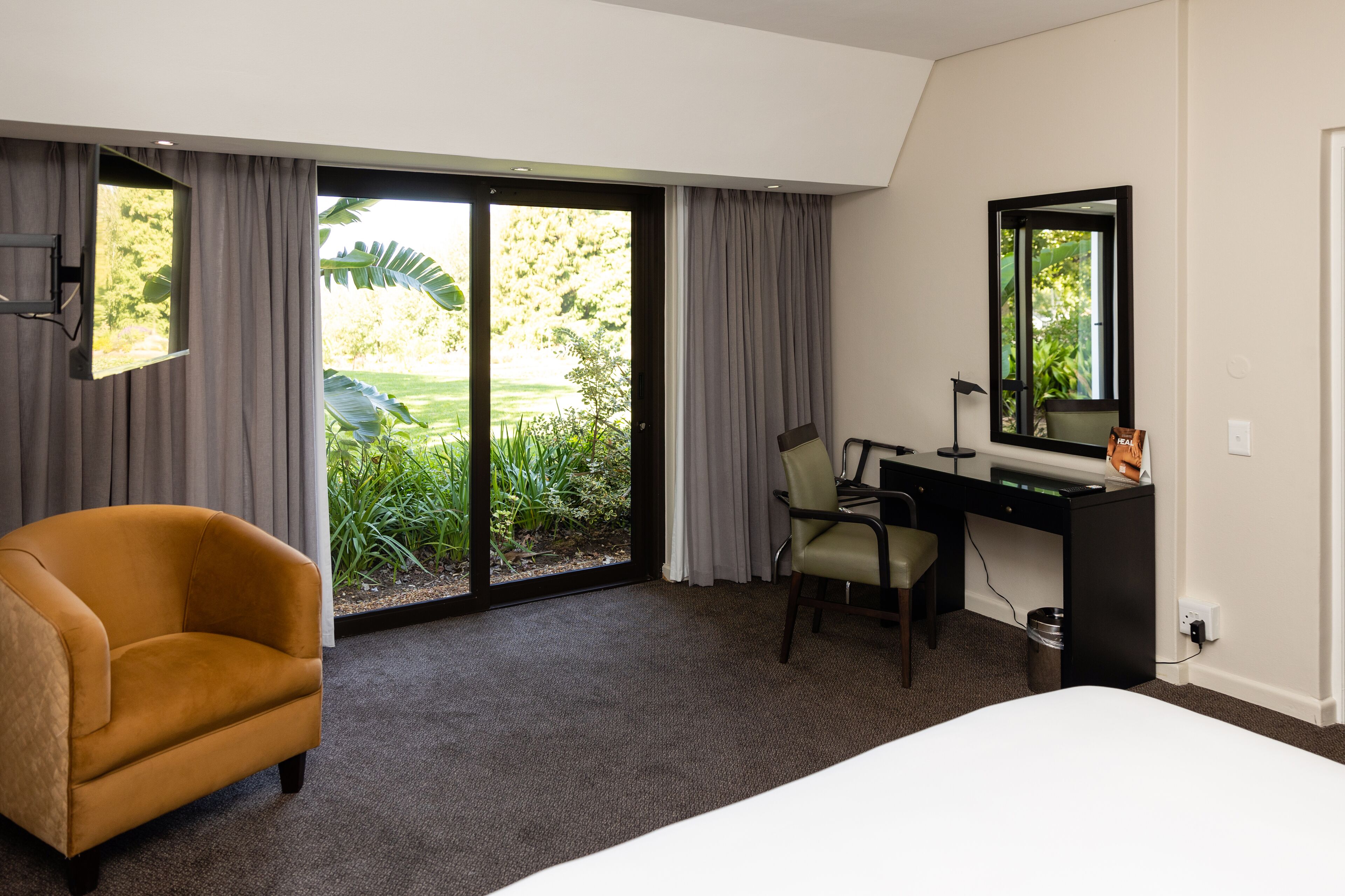 A hotel room with a large bed, a desk with a chair, a small armchair, and a sliding glass door leading to a garden.