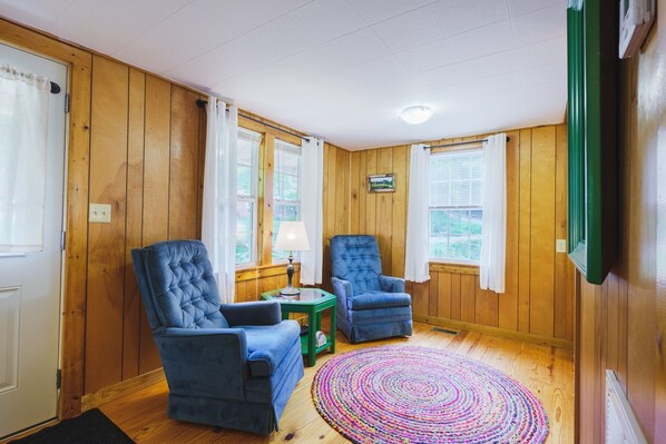 A cozy room with wooden walls and floors, two blue armchairs, a small table with a lamp, and a colorful circular rug.