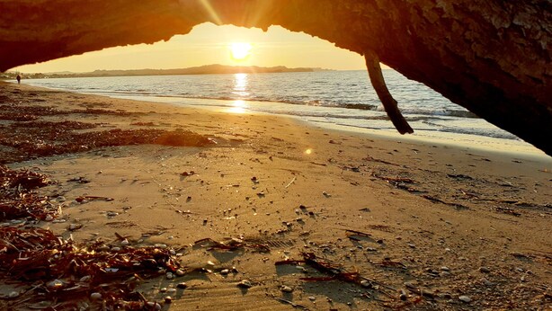 Escena de playa con una gran formación rocosa en primer plano y el sol poniéndose al fondo.