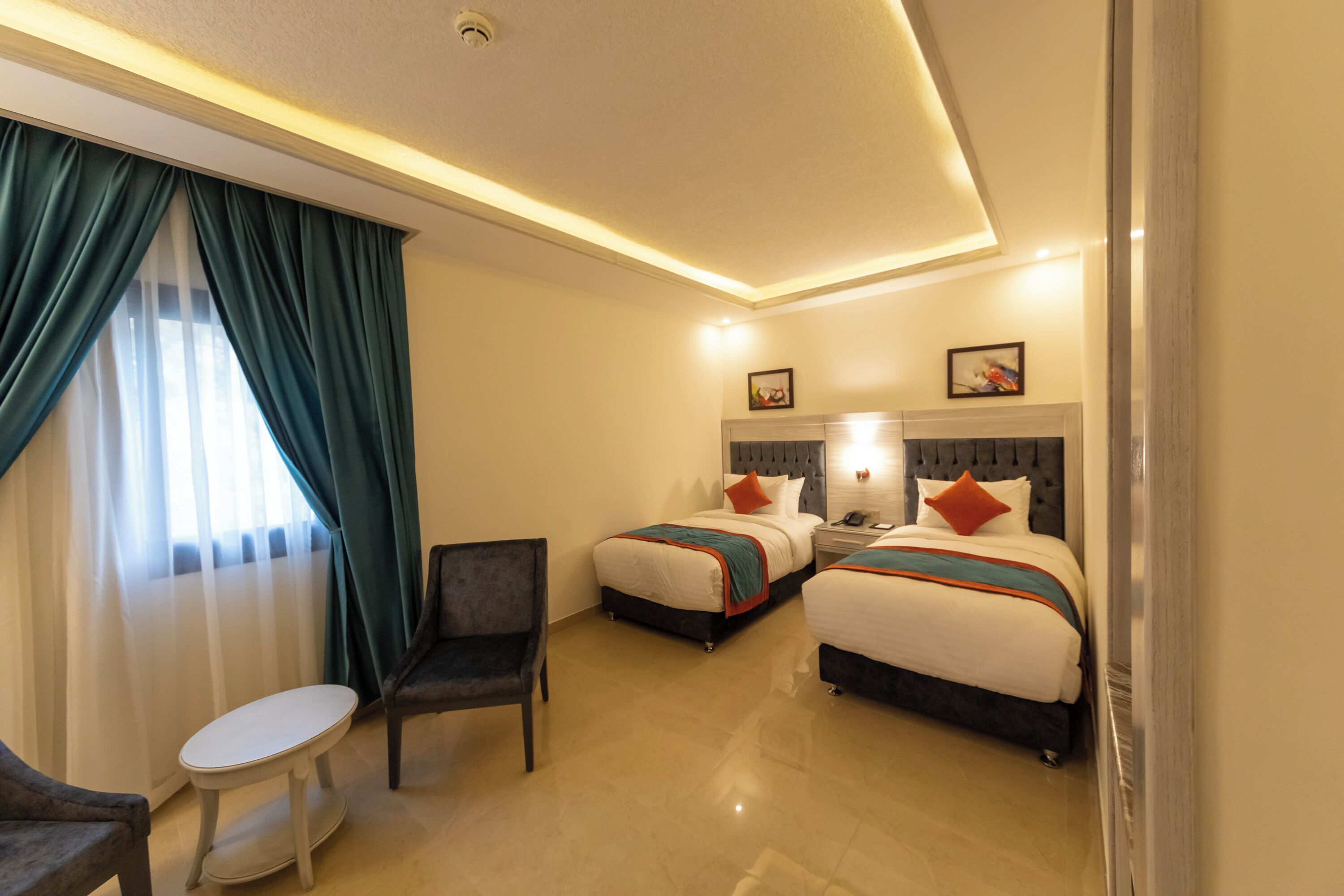 A hotel room with two beds, a small table, a chair, and a window with curtains.