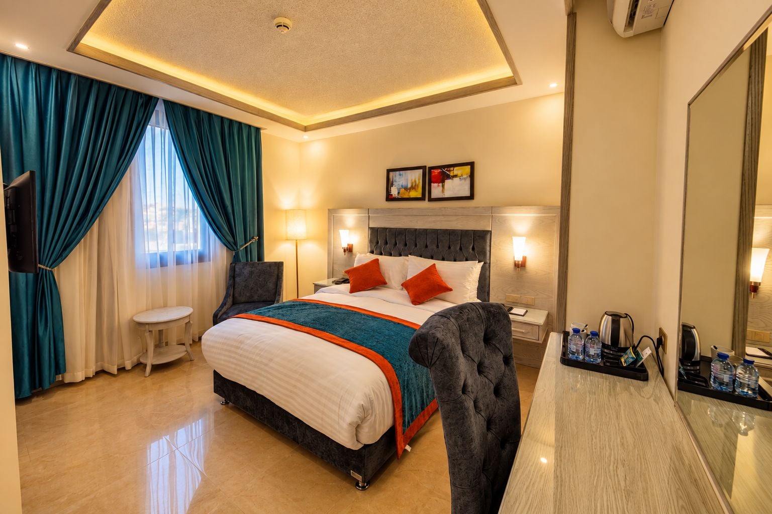 A hotel room with a large bed, a desk, a chair, a small table, a mirror, and a wall-mounted TV.