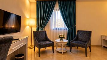 A hotel room with a flat-screen TV, two armchairs, a small round table with a vase of flowers, and a lamp.