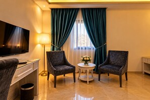 A hotel room with a flat-screen TV, two armchairs, a small round table with a vase of flowers, and a lamp. - Petra Canyon Hotel (Wadi Musa)