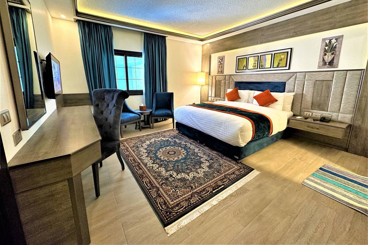 A hotel room with a large bed, a desk, a chair, a small table, and a large rug.