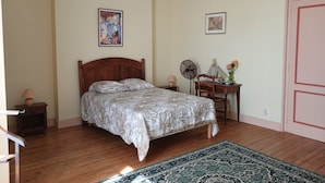 A bedroom with a wooden bed, a patterned bedspread, a standing fan, a small table with a lamp, and a door.