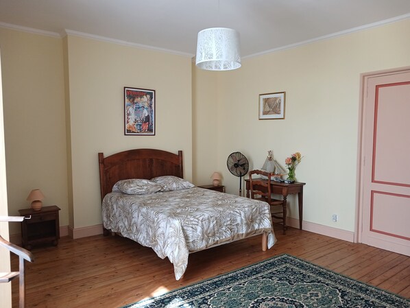 Une chambre avec un lit en bois, une couvre-lit à motifs, un ventilateur sur pied, une petite table avec une lampe et une porte.