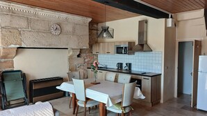 A cozy kitchen with a stone wall, wooden ceiling beams, a dining table with chairs, a microwave, and a refrigerator.