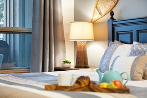 A neatly made bed with a tray of breakfast items, a bedside lamp, and a view of the outdoors through a window with curtains.