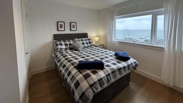 A bedroom with a checkered bedspread, a bedside lamp, a window with a view of the sea, and a hanging light fixture. - Carleys House (Macduff)