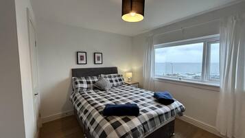 A bedroom with a checkered bedspread, a bedside lamp, a window with a view of the sea, and a hanging light fixture.