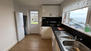 A compact kitchen with a built-in washing machine, a large window with a view of the sea, and a door leading outside. - Carleys House (Macduff)
