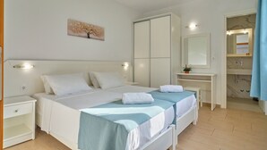Een tweepersoonsbed met witte en blauwe beddengoed, een nachtkastje, een bijzettafeltje, een spiegel en een kleine potplant.