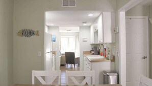 Cocina con gabinetes blancos, un lavamanos y un cubo de basura. En primer plano, se observa una mesa de comedor con sillas.