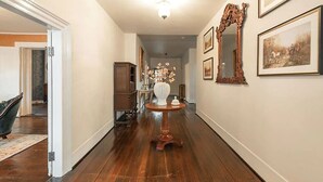 Un pasillo con suelo de madera, una puerta blanca, una mesa de estilo antiguo con un jarrón y cuadros en las paredes.