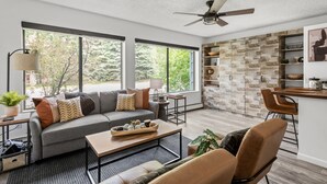 A modern living room with a sofa, coffee table, and chairs, a kitchen area with a dining table, and large windows offering a view of trees. - HOLT - Billionaire Mountain (Aspen)