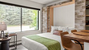 A modern hotel room with a large window, a bed with a green throw, a wooden dining table, and a built-in wardrobe. - HOLT - Billionaire Mountain (Aspen)