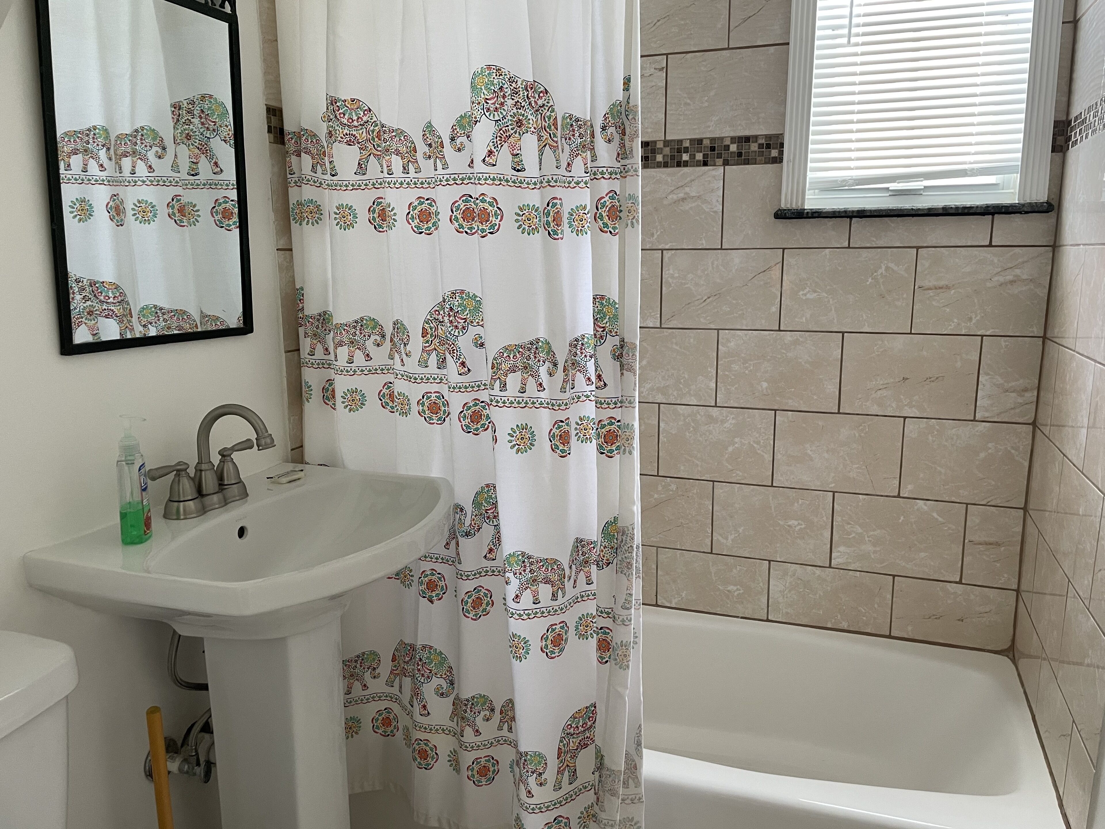 Une salle de bain avec un lavabo blanc, un miroir, un rideau de douche orné de motifs d’éléphants et des murs recouverts de carreaux beiges.