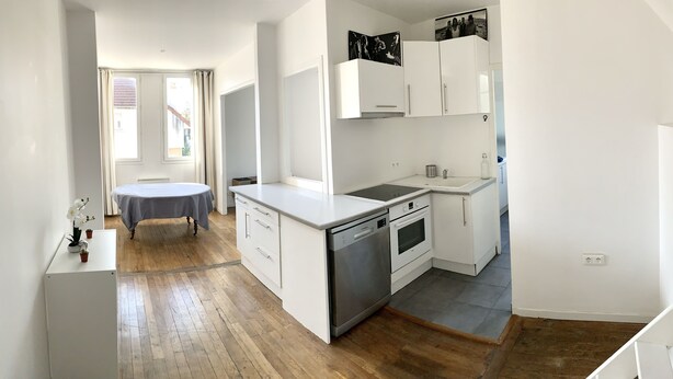 A compact kitchen with white cabinetry, a dishwasher, an oven, and a sink. There is a small dining table with a chair, and a window with curtains. - House (Arcueil)