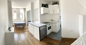 A compact kitchen with white cabinetry, a dishwasher, an oven, and a sink. There is a small dining table with a chair, and a window with curtains.
