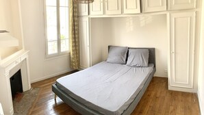 A bedroom with a bed, wooden flooring, a window with curtains, and white cabinets above the bed. - House (Arcueil)