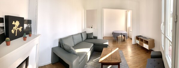 A modern living room with a grey sectional sofa, a wooden coffee table, and a small round table with a grey tablecloth. There is a bookshelf and a window with a view of the outside.