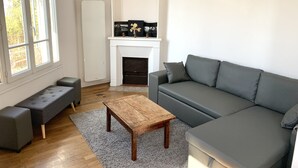 A modern living room with a grey sofa, a wooden coffee table, a grey bench, and a fireplace with decorative items. - House (Arcueil)