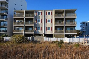 Un immeuble résidentiel à plusieurs étages, doté de balcons, d’une clôture blanche et d’une pelouse sèche au premier plan.