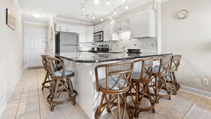 Une cuisine avec des meubles blancs, un îlot central doté de tabourets, un réfrigérateur et un four à micro-ondes.