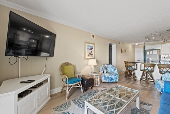 A living room with a flat-screen TV, wicker furniture, a patterned rug, and a kitchen area with bar stools.