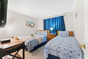 A hotel room with two beds, a desk, a TV, and a window with blue curtains.