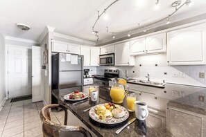 Une cuisine moderne avec des meubles blancs, un réfrigérateur en acier inoxydable, un four à micro-ondes et un grand îlot central où sont disposés des aliments pour le petit-déjeuner.