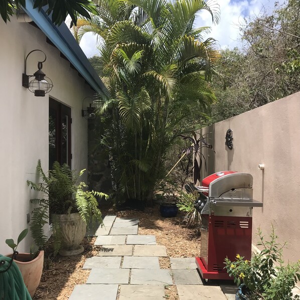 A narrow stone pathway leading to a house with a red barbecue grill, surrounded by lush greenery and a blue roof. - BLUE KAI, Water View, 2 King Suites Outdoor Native Stone Showers (St John)