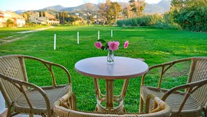 Mesa de mimbre con dos sillas y un jarrón de flores en un espacio al aire libre con césped y vistas a casas y montañas al fondo.