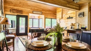 Interior de madera con una mesa de comedor para cuatro personas, un sofá y una zona de cocina con un reloj y una planta.