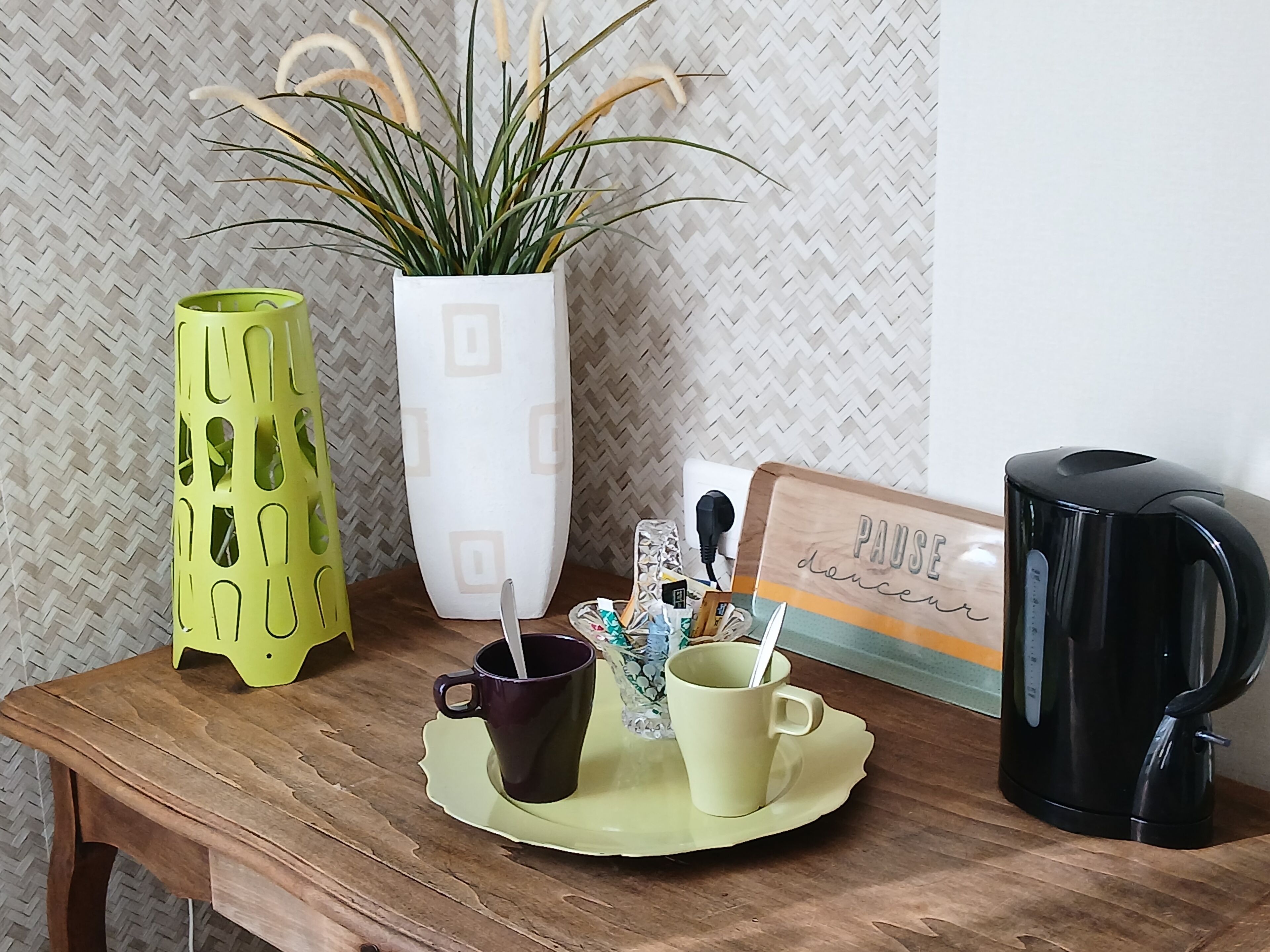 Una mesa de madera con una planta en maceta, un jarrón verde, una cafetera y una bandeja con dos tazas.